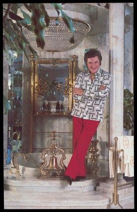 Liberace, wearing red trousers. Colour photographic postcard after A. Leibowitz, 1981.