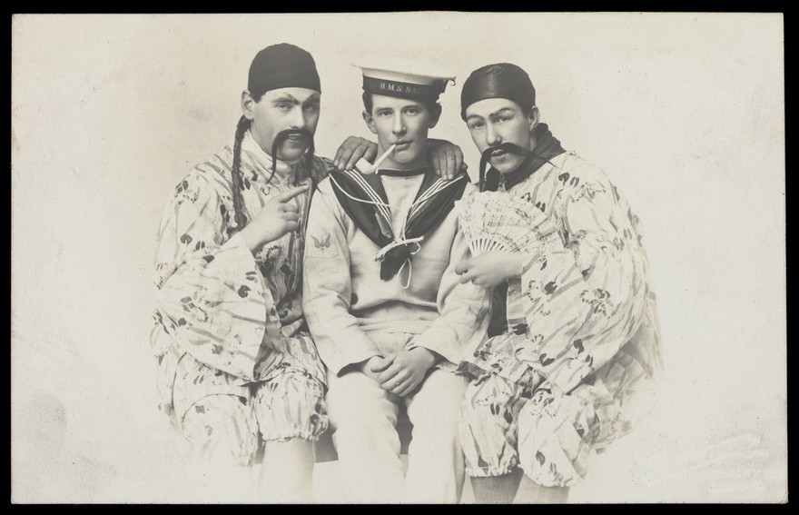 Three sailors from H.M.S. Natal sitting together: one is in uniform, the other two are wearing pseudo-Oriental costumes. Photographic postcard by T.E. Lonnergan, 191-.