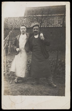 Two men, standing together in a garden, one with his arm around the other. Photographic postcard, 1907.