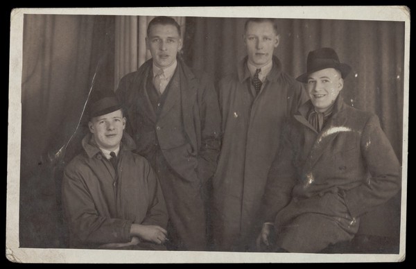 Four men posing together wearing street clothes and coats, in front of a curtain. Photographic postcard, 194-.