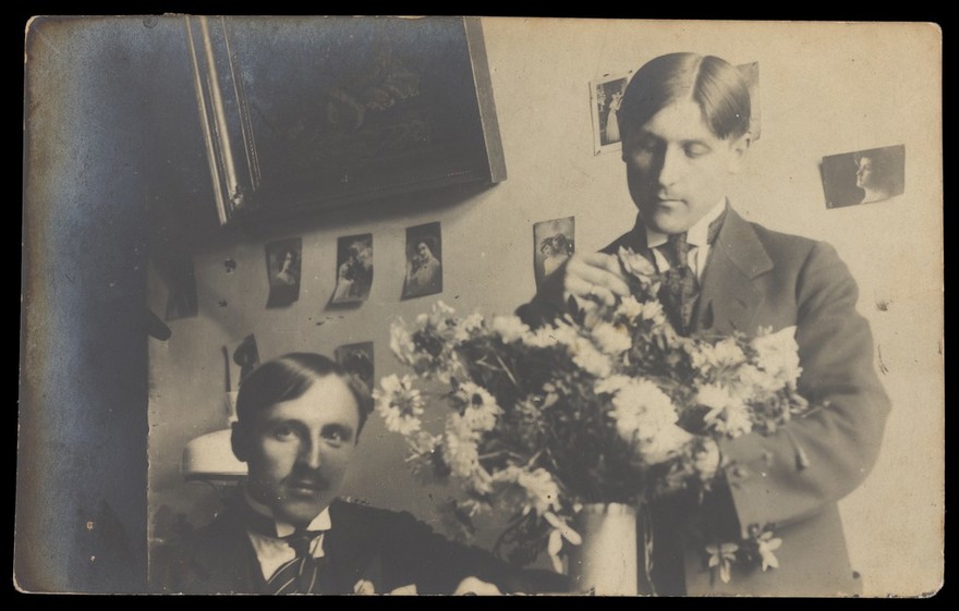 Two men in a domestic setting, one of them arranging flowers. Photographic postcard, ca. 1910.