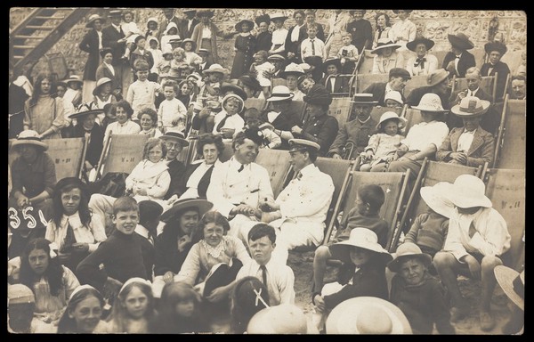 Families sitting in deckchairs, with two men in naval uniforms embracing in the centre. Photographic postcard, 191-.
