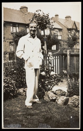 A man poses wearing a white suit in a garden. Photographic postcard by Jerome Ltd., 192-.