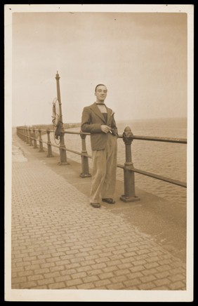 A man with a cigarette posing on a promenade by the sea. Colour photographic postcard by Jerome Ltd., 192-.