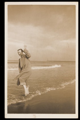 A man in a long coat paddling in the sea. Colour photographic postcard, 192-.