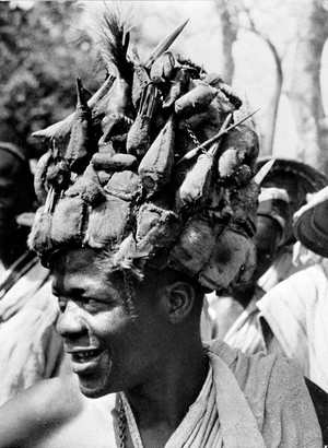 view A West African witchdoctor with a hat entirely covered with amulets of leather and feathers. The leather is sewn into little pouches, each of which contains verses from the Koran and other charms.