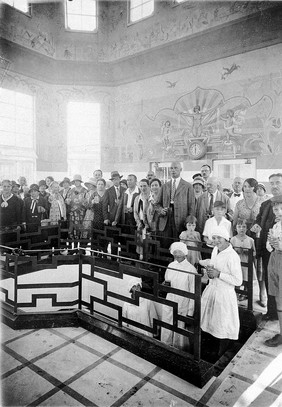 Franzensbad (Františkovy Lázně), Czechoslovakia: the interior of the Natalie Spring where people stand to drink water carried up steps from a central point by female attendants wearing white uniforms and caps. Photograph.