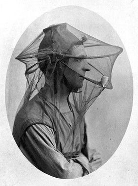 Mosquito headnet with a cap, modelled by a man in profile smoking a pipe. Photograph, 1902/1918 (?).