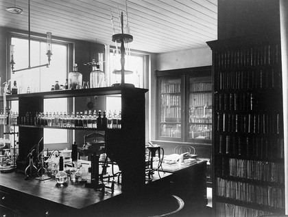 The interior of a pharmaceutical or chemical laboratory with a bench in the centre and books and papers on shelves around the walls. Photograph.