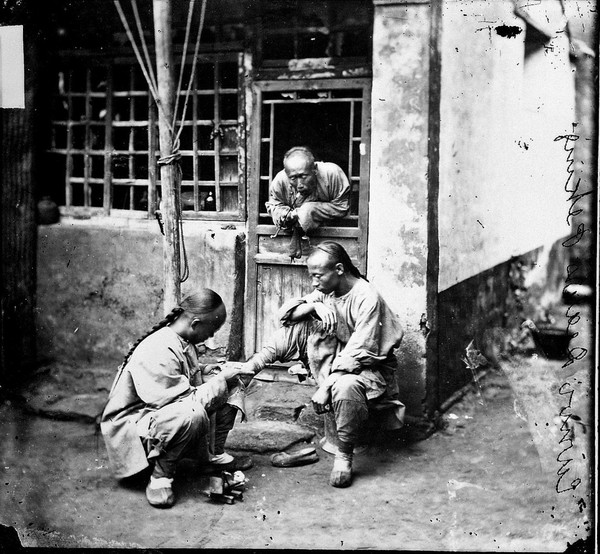 A travelling chiropodist operating upon a corn on his patient's foot. Peking, Pechili province, China.