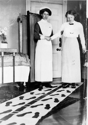 A nurse steadying a woman by holding her right arm, while the woman attempts to walk along a mat marked with footprints. Photograph.