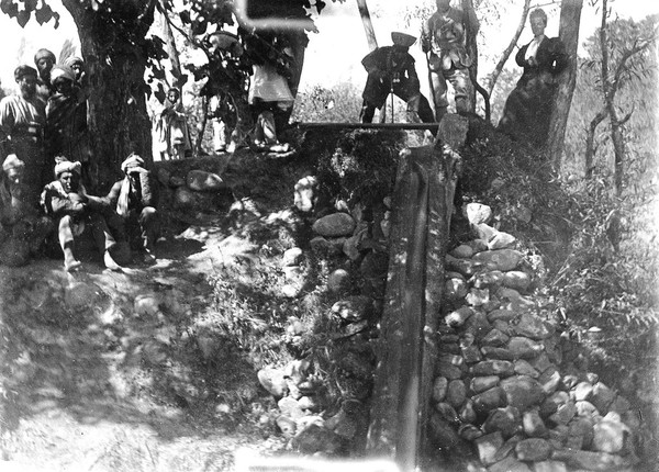 The Indian Punjabi Mir Munshi Sultan Mohammad standing, legs astraddled, over a water channel. Next to him is a European Lady, possibly the nurse Mrs. Kate Daly. Kabul