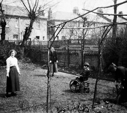 A physically disabled boy sitting in a wheelchair in the back garden of a semidetached house. Photograph, ca. 1910/1925.