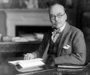 view Portrait of Ronald Ross, seated at a desk wearing glasses, with a pen in his hand.