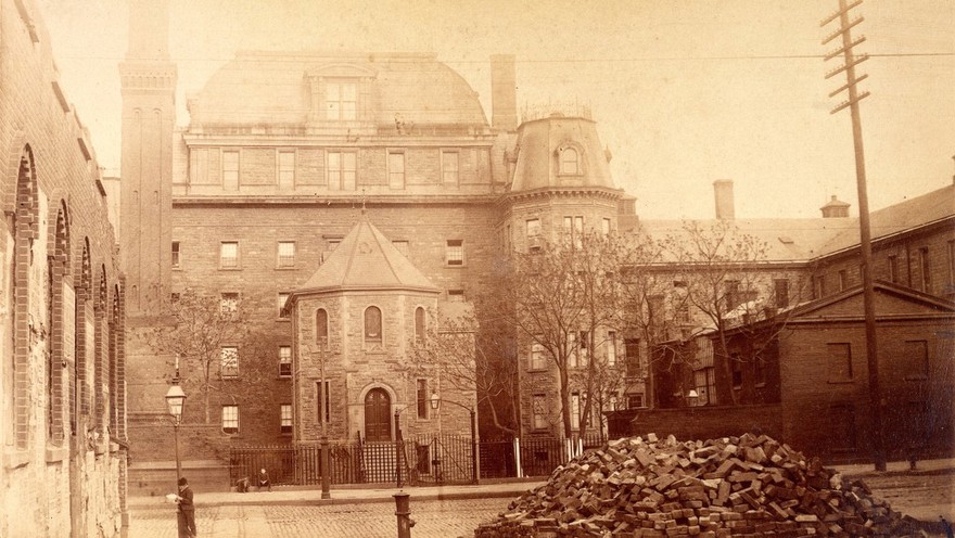 Bellevue Hospital, New York City: hospital buildings with a tower; a pile of bricks in the foreground from a demolished building. Photograph.