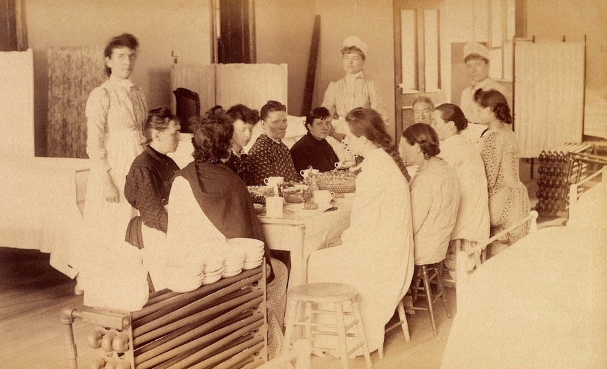 Bellevue Hospital, New York City: women patients (mentally ill?) having a meal in a ward, with three nurses. Photograph.