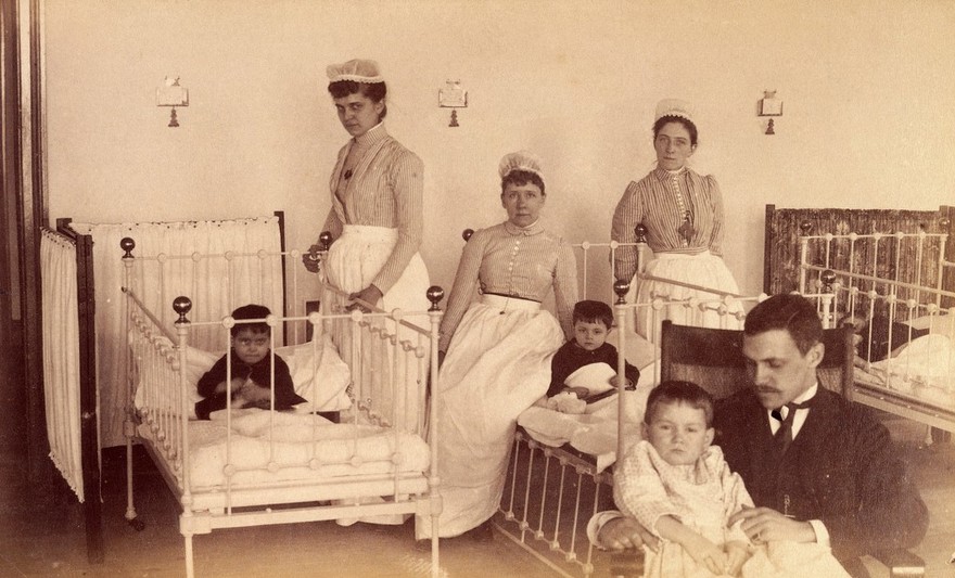 Bellevue Hospital, New York City: children in a ward with nurses and a doctor (?). Photograph.