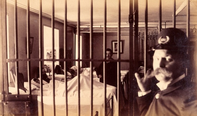 Bellevue Hospital, New York City: male patients (criminal insane?) in bed in ward, prison bars in foreground guarded by policeman. Photograph.