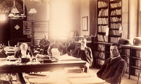 Bellevue Hospital, New York City: a library with patients(?), doctors (?), possibly patient ledgers in bookcase. Photograph.