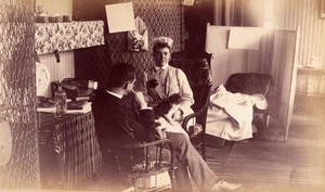 view Bellevue Hospital, New York City: a nurse's sitting room (?), or end of ward, with seated nurse, cat in lap, talking to seated man. Photograph.