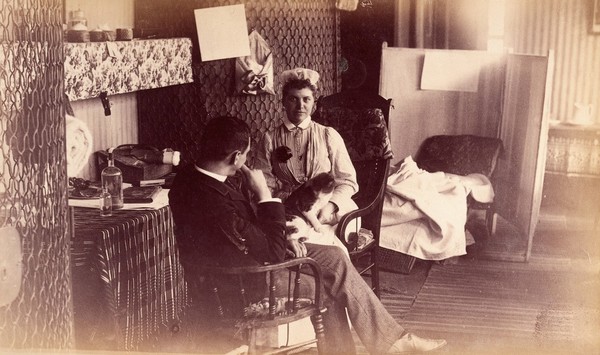 Bellevue Hospital, New York City: a nurse's sitting room (?), or end of ward, with seated nurse, cat in lap, talking to seated man. Photograph.