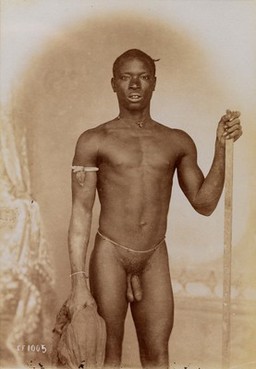 A young African man posing naked, holding a stick and a piece of cloth, in front of a painted backdrop. 