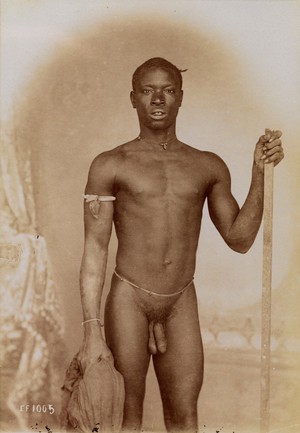 view A young African man posing naked, holding a stick and a piece of cloth, in front of a painted backdrop.