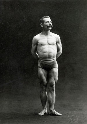 view A male bodybuilder wearing bathing trunks, posing in a studio setting.