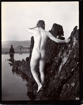 A man posing naked, with his back to the camera, standing on a rocky outcrop.