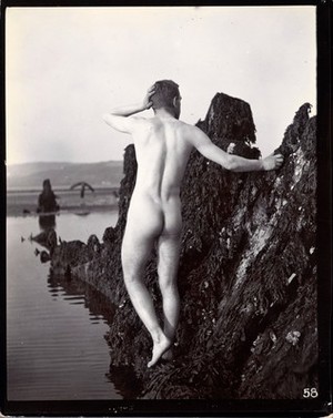 view A man posing naked, with his back to the camera, standing on a rocky outcrop.