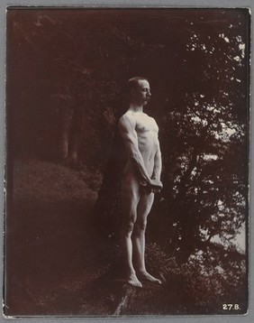 A man posing naked, standing with his chest out, arms down and hands clasped.