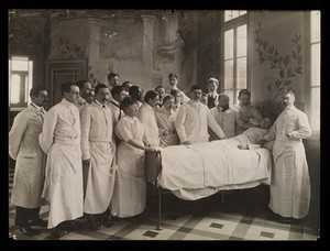 view Medical staff standing round a woman patient in bed in a hospital ward. Photograph by Seeberger Frères, ca. 1910.