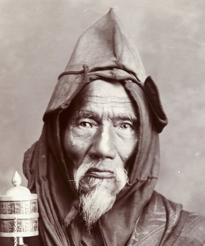 A bearded elderly man, carrying a prayer wheel and a rosary, in a studio setting.
