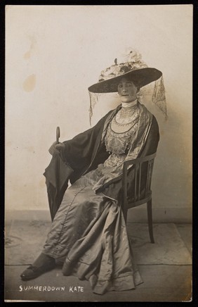 A convalescent soldier (Lance-Corporal R.C. Thomas) poses in drag, wearing a silk dress and sitting with a mourning veil. Photographic postcard, 1916.