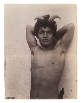 A Sicilian boy naked from the waist up, his hands behind his head, wearing horns.