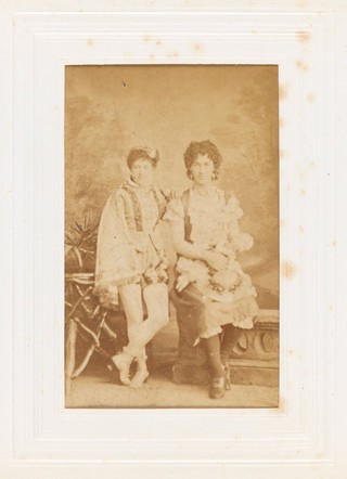 Two people in theatrical costume side by side, in front of a painted backrop.