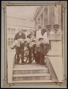 Four men wearing aprons and uniforms standing by a trolley with half-dissected cadavers on it. Photograph.