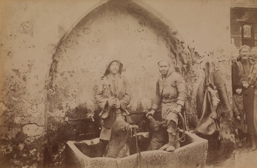 Santorini, Greece: men carrying hogskins full of water to a cistern. Photograph. 18--.
