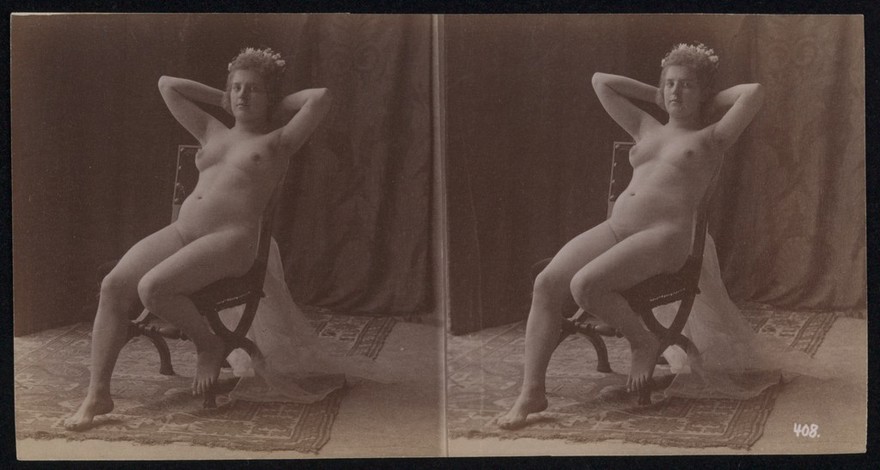 A young woman posing naked but for a long veil, leaning back in a chair, in a photographic studio, in front of a curtain. Stereo photograph, ca.1900.