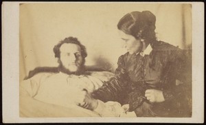 view A bearded man lying in bed, his hand held by his wife who is seated next to the bed. Photograph, 187-.
