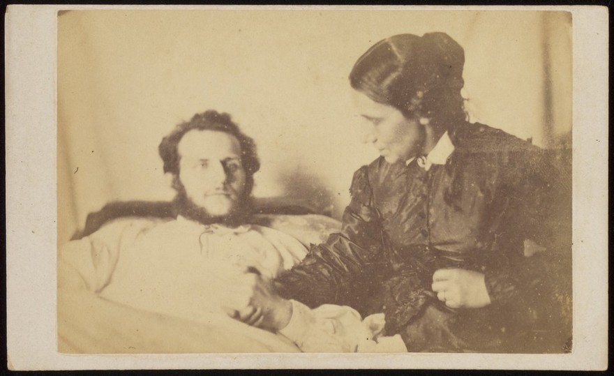 A bearded man lying in bed, his hand held by his wife who is seated next to the bed. Photograph, 187-.