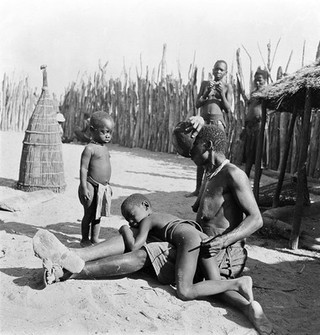 Angola: a Kwanyama woman performing an enema on a small child: the woman takes liquid into her mouth from a bowl. Photograph by Antoinette Powell-Cotton, 1936/1937.