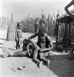 view Angola: a Kwanyama woman performing an enema on a small child: with the child placed across her lap the woman prepares a tube. Photograph by Antoinette Powell-Cotton, 1936/1937.