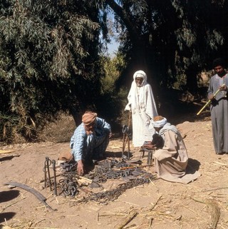 Luxor, Egypt; blacksmith selling his wares. Shows men, one with a goat, examining metal implements. Trades such as smithing are often performed by landless villagers who are often amongst the poorest groups. In 1990, as much as 40% of the rural population was landless. These people often provide village services including carpentry, machinery maintenance, and livestock herding or cultivate land for absentee landlords as tenants and sharecroppers. Photographed in January 1990.