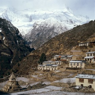 Nepal; town life in the Khumbu, 1986. Namche Bazar (altitude 3446 metres), a bustling and prosperous market town, is the largest Sherpa settlement in the Khumbu. Many of its Tibetan-style houses devote the ground floor to animal shelters while the family lives upstairs. Most have windows and doors at the front of the building only, the back being built into the side of the mountain. Firewood is stacked against walls, and small terraced fields grow staples (potatoes, barley, wheat). At left is a Buddhist shrine or stupa, on each side of which is painted the eyes of the Buddha. Prayer flags are strung out from its summit.