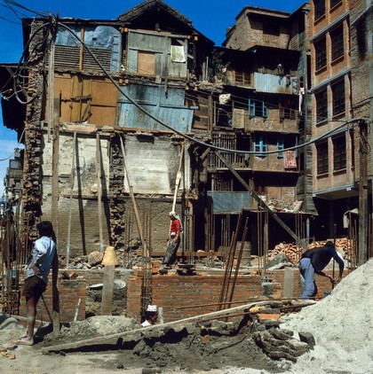 Nepal; tenements, old and new, Kathmandu, 1986. Builders erecting new apartments on the site of an old tenement block. Once erected, the residents of the existing four-storey tenement block will have their daylight extinguished. A woman picks her way across the building site from her home to the street.