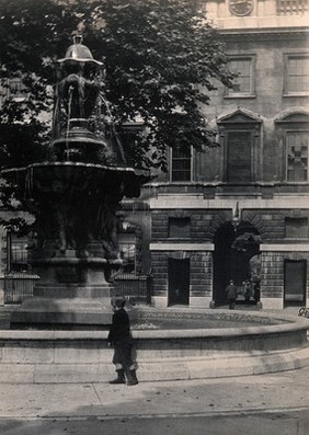 St Bartholomew's Hospital, London: the fountain in the centre of the courtyard with a small boy looking at it, in the background the gateway to Smithfield. Photograph.