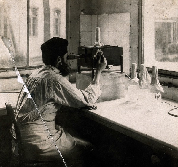 A man controlling the titration of bacteria in a vaccine with the aid of an optical apparatus. Photograph, [ca. 1940].