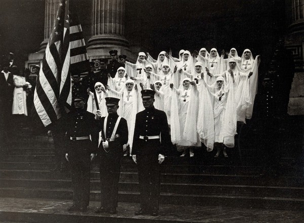 World War One nurses, with arms outstretched, stand with soldiers on the steps of a large building during a ceremonial occasion, New York City. Photograph by Underwood & Underwood, 1918.