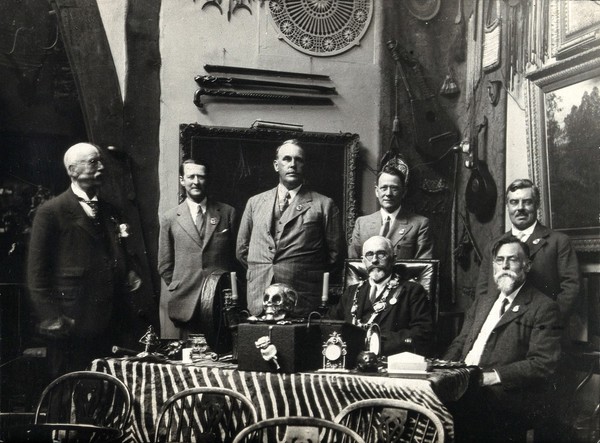 A museum interior (?): seven men pose with collected objects, including a human skull, displayed on a table. Photograph, ca. 1930.